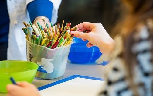 Person chooses a paintbrush from a container; art supplies and bowls are on the table.
