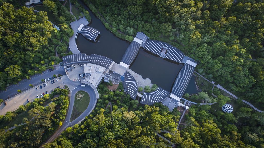 Aerial view of modern buildings with curved roofs among trees and connected by water.