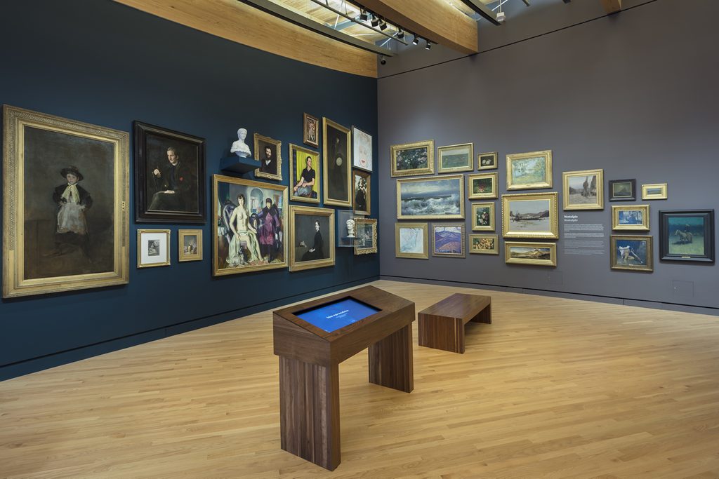 Museum gallery with framed artworks, digital table, and wooden bench on light wood floor.