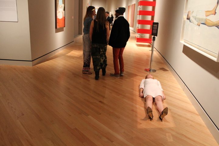 An actor plays Supine Woman Art gallery with people observing, woman lies on floor, paintings displayed on walls.