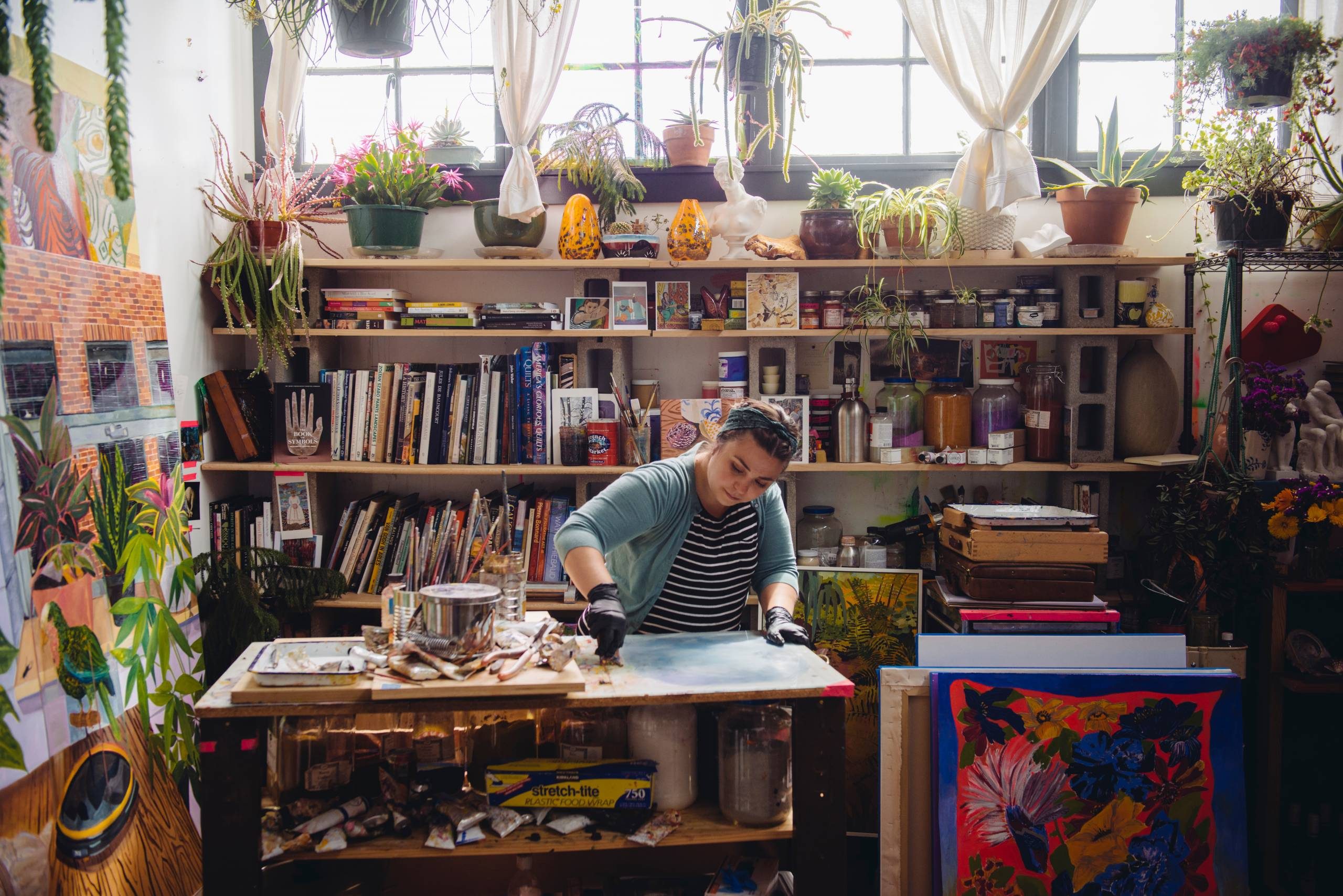 Artist working at a cluttered studio table with books, art supplies, plants, and artwork.