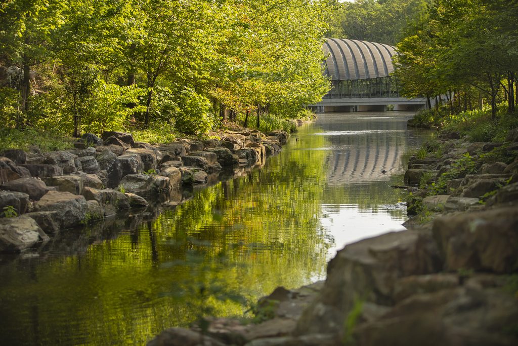 A pond with rocks and trees reflects a glass-front structure in a serene, natural setting.