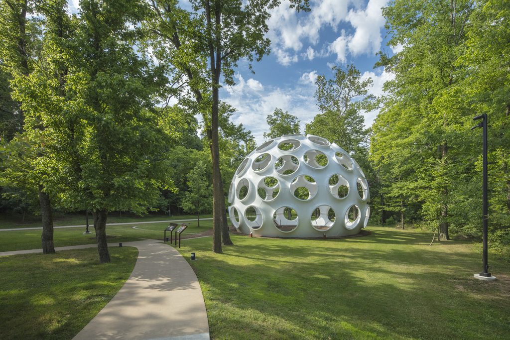 Geodesic dome with circular openings in a green park, trees, path, partly cloudy sky.