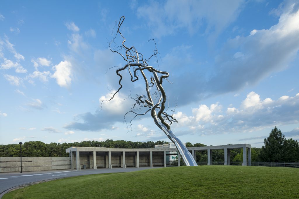 Abstract metallic tree sculpture on grass, modern building in background, cloudy sky above.