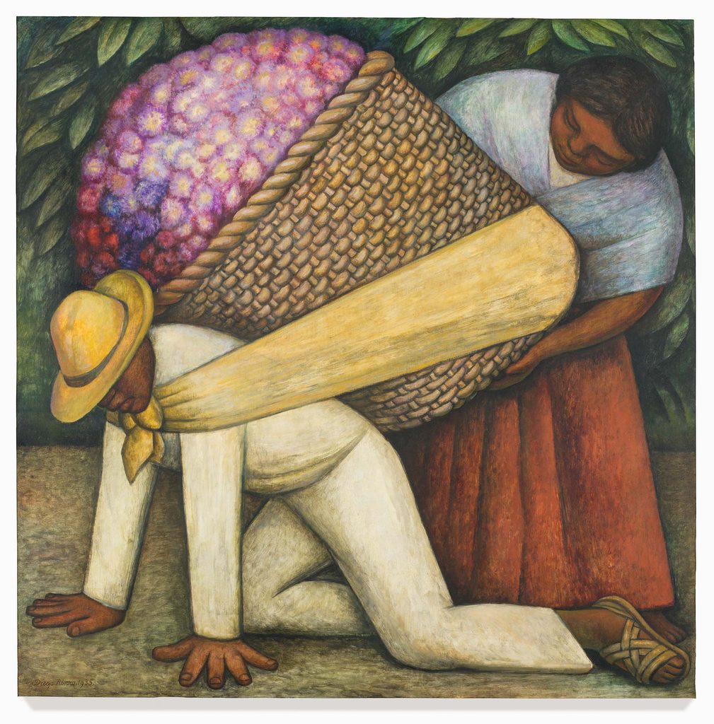 Man on ground with large basket of flowers tied to his back and a woman helps him balance it