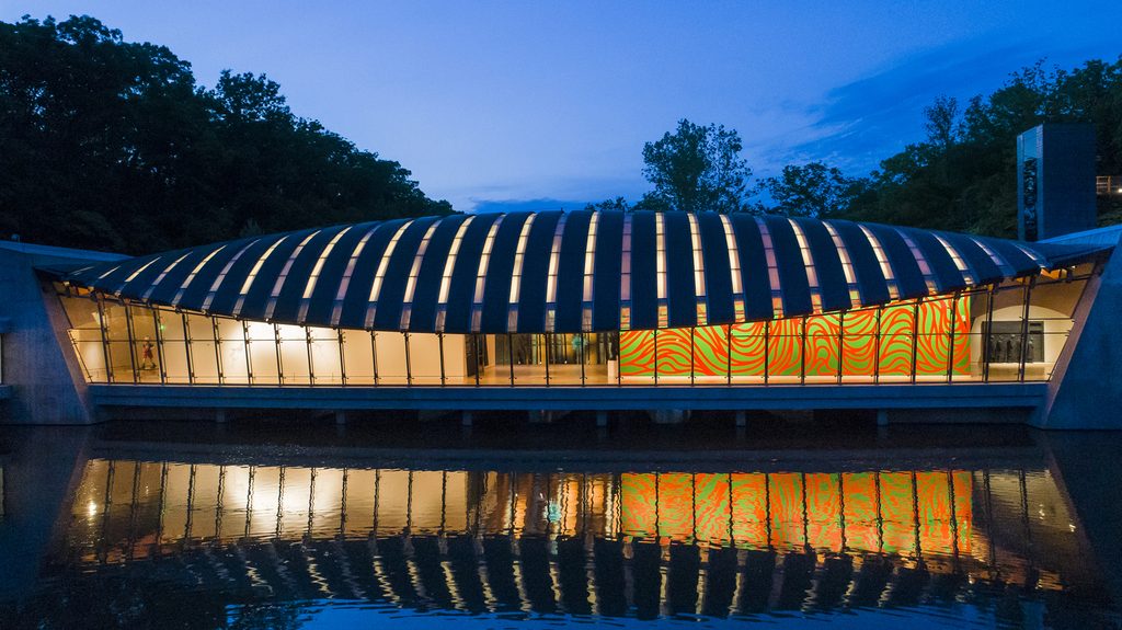 Modern building with curved roof, large windows, colorful pattern, reflecting on water at dusk.