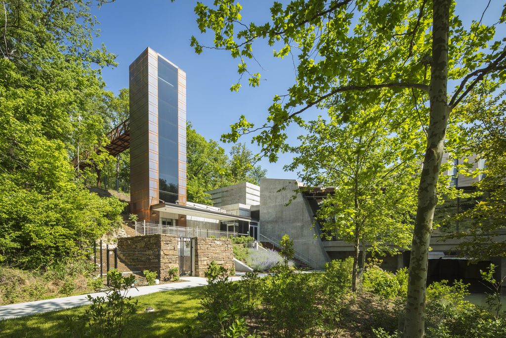 Modern building with glass tower in lush wooded area, featuring concrete elements and trees.