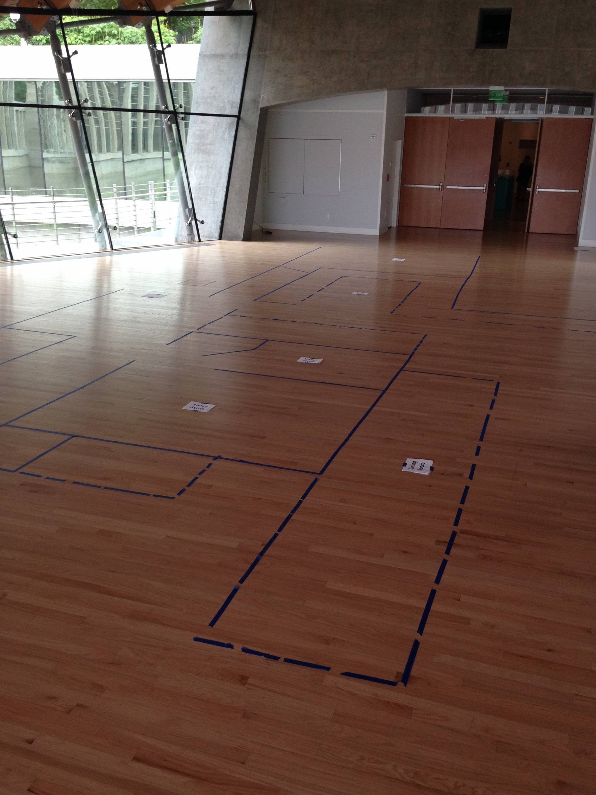 Spacious room with taped outlines on wooden floor, large windows, and double doors in background.