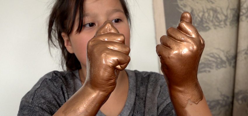 Person with metallic bronze-painted hands raised indoors, wearing a gray top, neutral background.