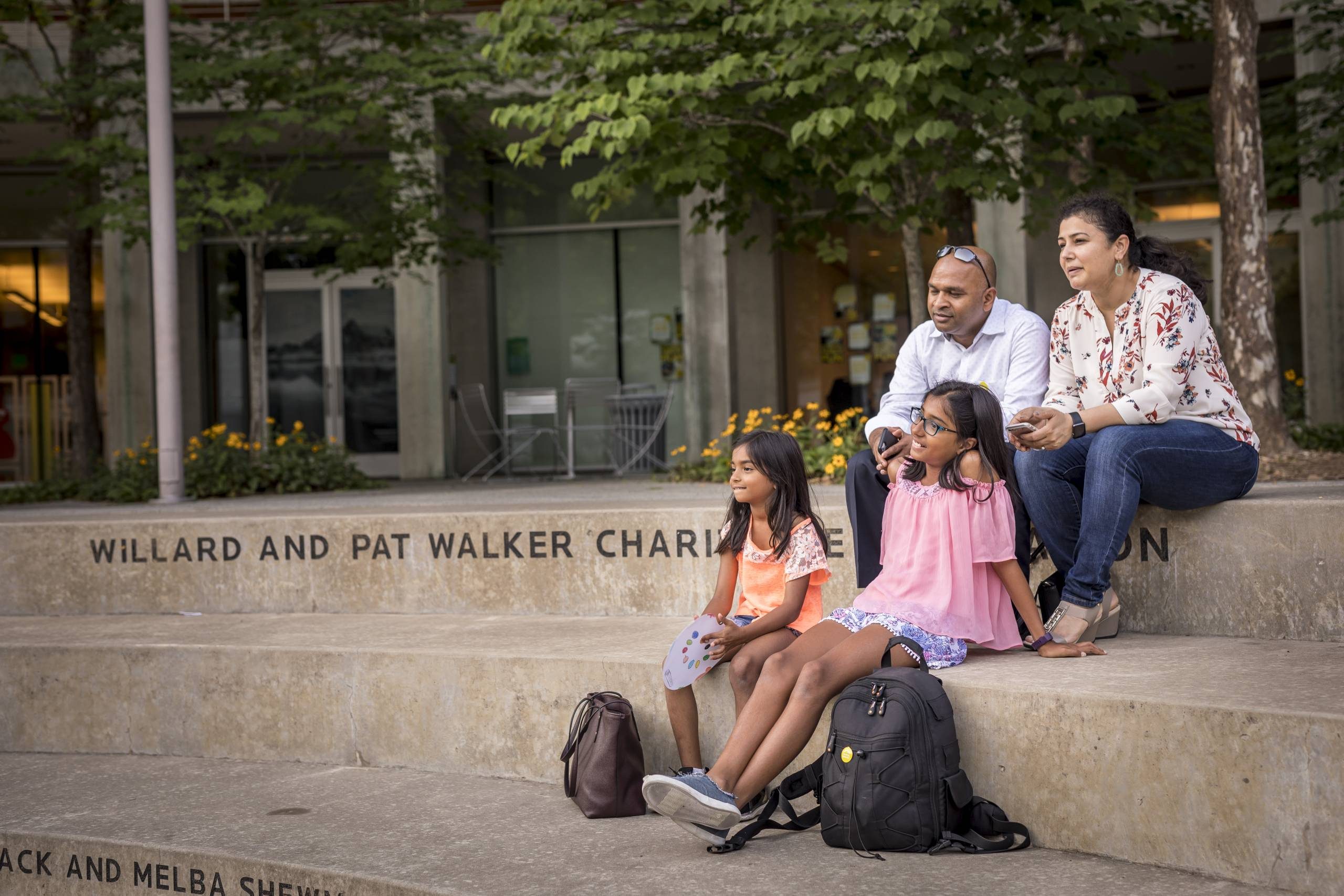 Family at Crystal Bridges Family of four seated on steps outdoors, with backpacks, near a glass-windowed building.