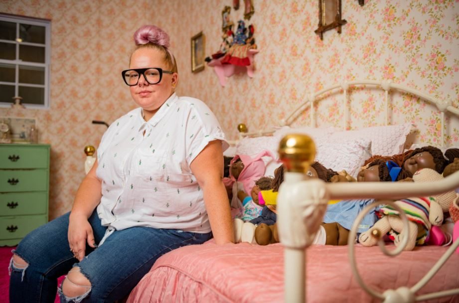 Genevieve Gaignard Person with topknot and glasses on vintage bed with dolls, pink bedspread, floral wallpaper.