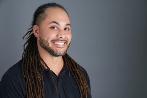 A smiling person with long hair and a beard in a dark shirt on a gray background.