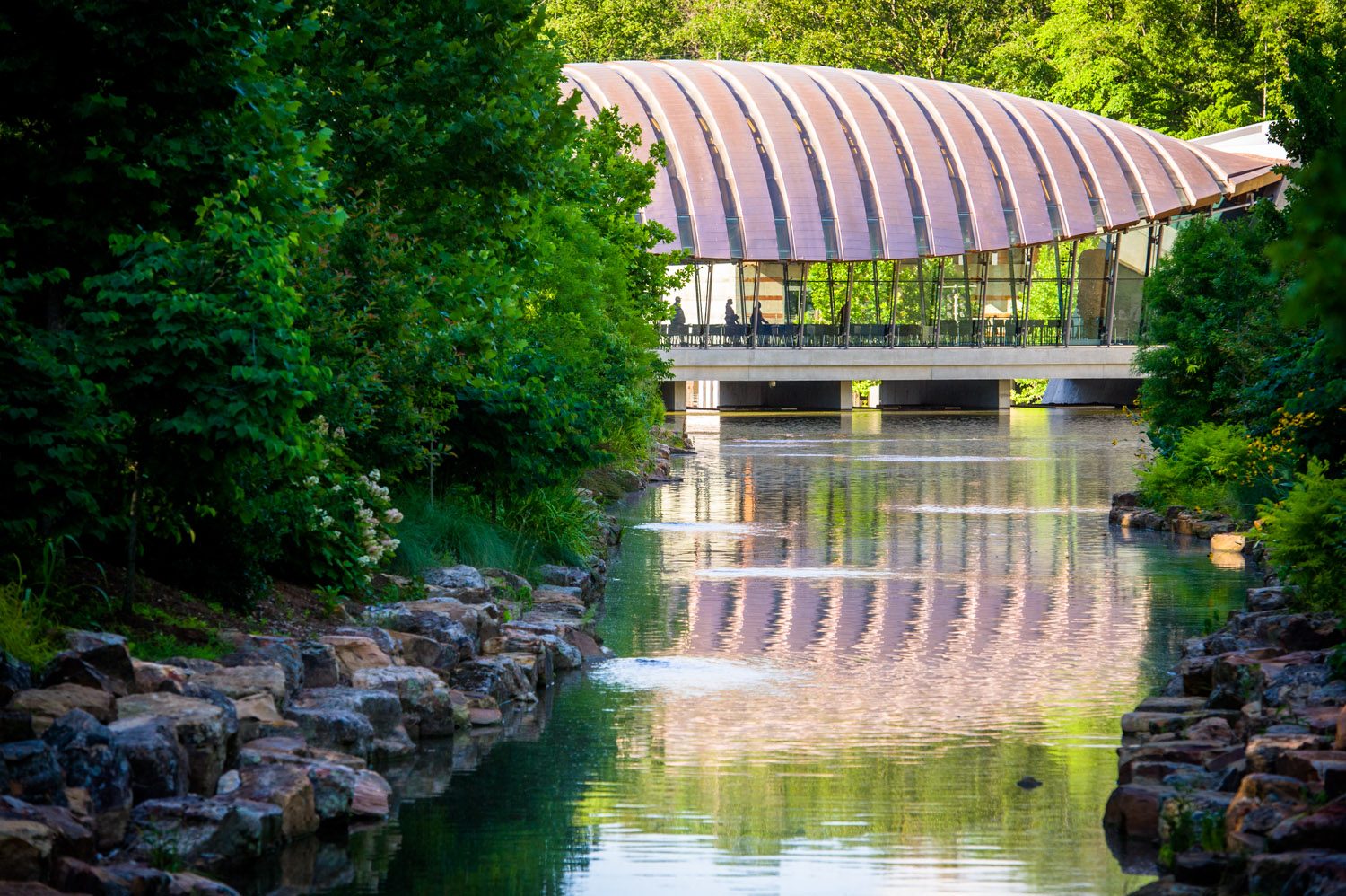 Summer at Crystal Bridges Modern curved-roof building over water, with lush greenery and stone-lined banks.