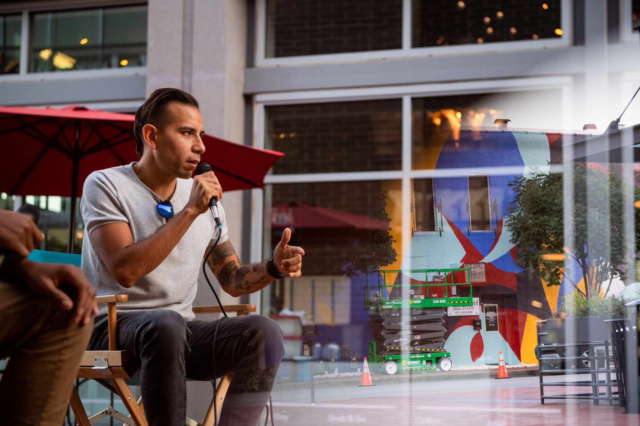 Person in a chair speaks into a microphone outdoors, with building and mural in the background.