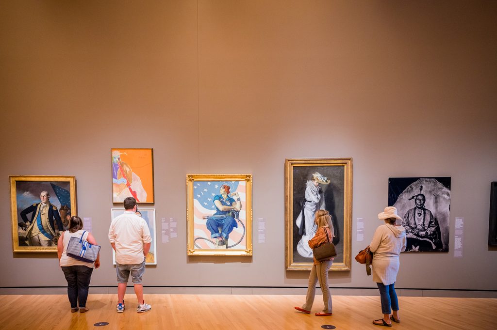 People viewing various paintings in a well-lit art gallery.