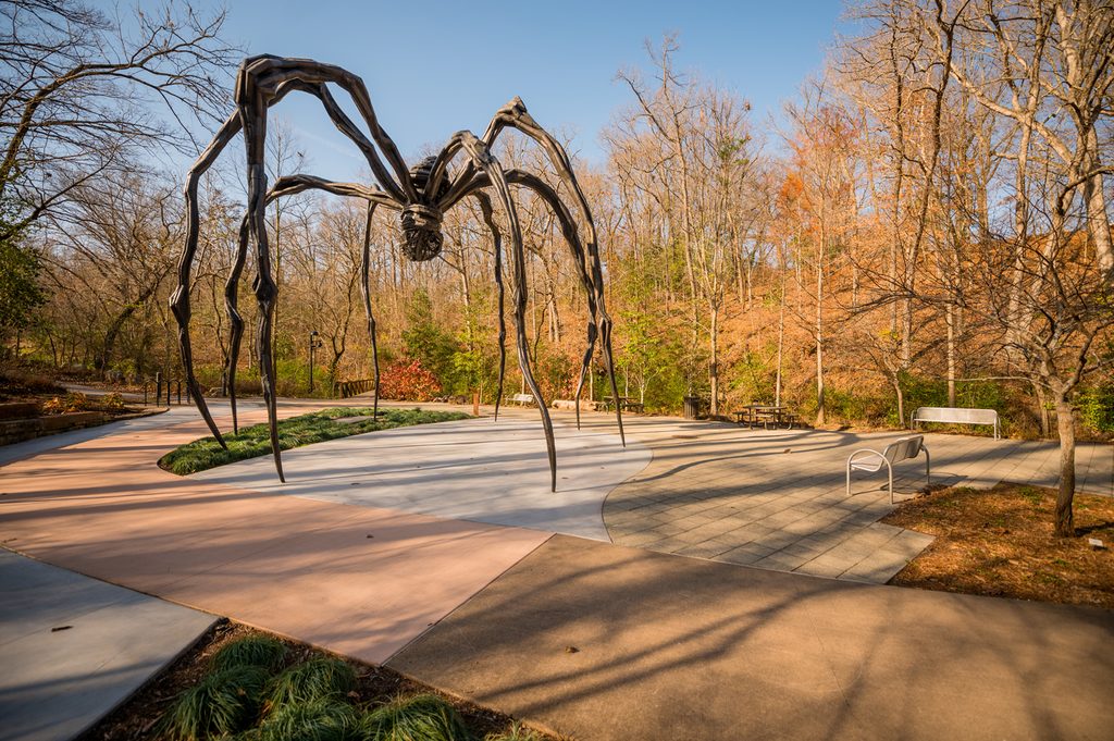 Metal spider sculpture in park with autumn trees, benches, and paved paths.