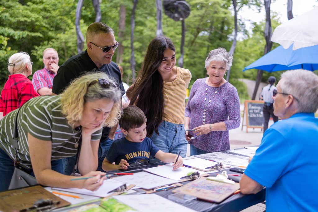 Group drawing and writing at a table outdoors in a park with greenery in the background.
