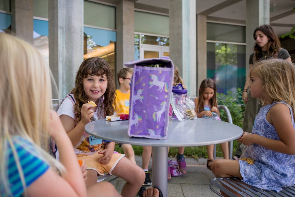 Children sitting at outdoor table having snacks, one girl smiling, unicorn lunch bag visible.