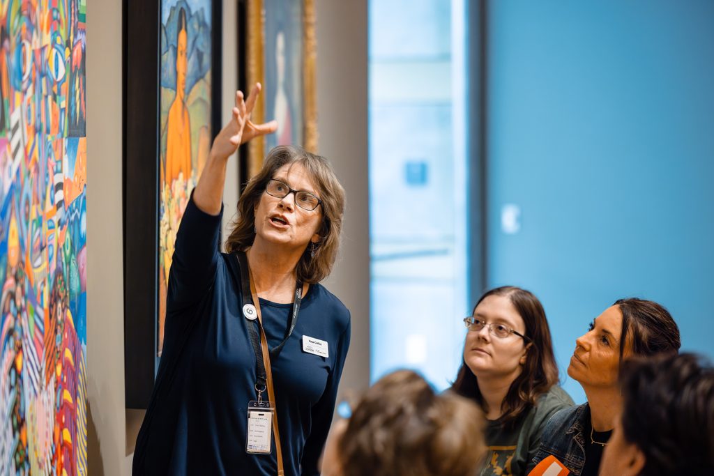 Woman presents colorful paintings in a gallery as people listen attentively.