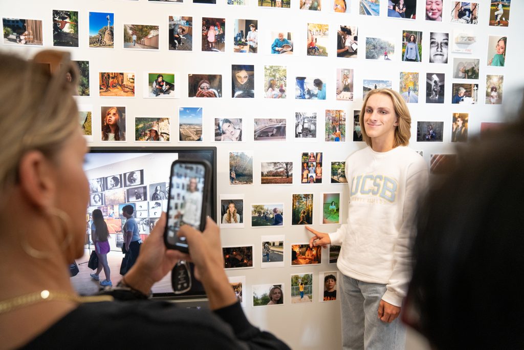 A teen photographer stands in front of a wall of photos that includes their work at the Through the Lens celebration at Crystal Bridges