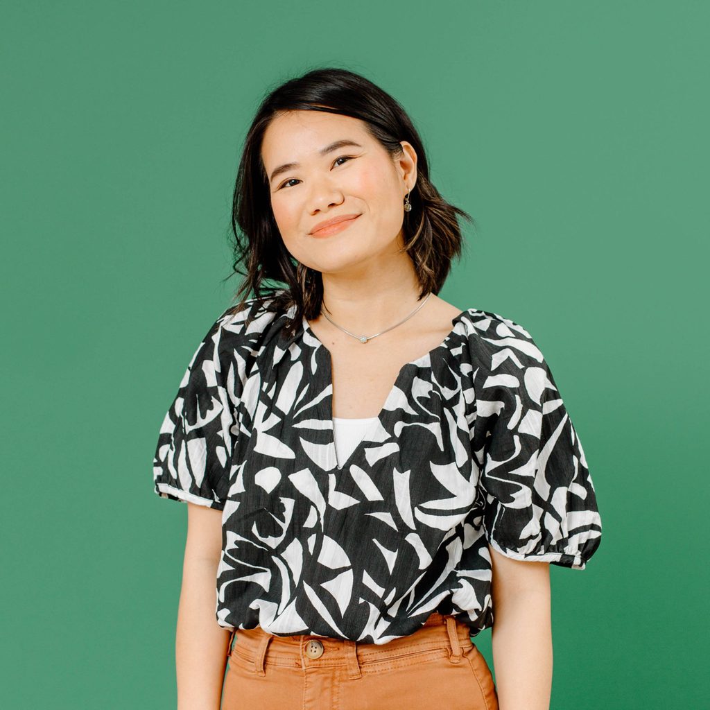 Person with short black hair in a patterned top and brown pants against green background.