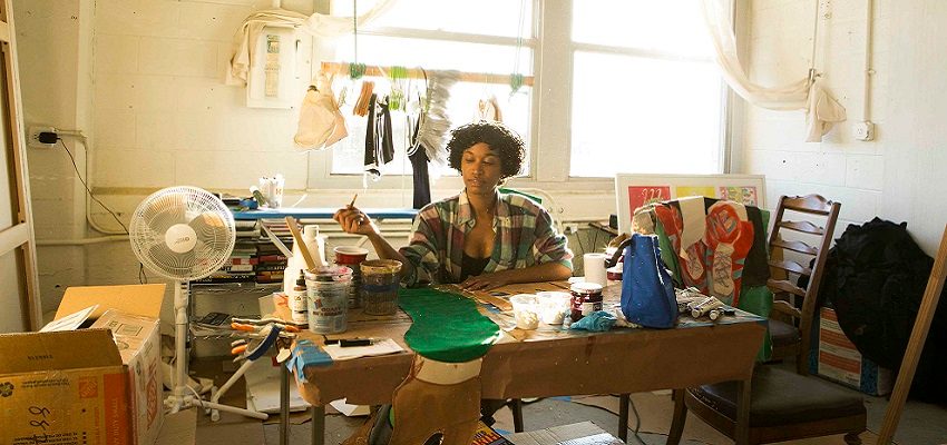 Artist painting at a cluttered table in a sunlit studio with art supplies and boxes visible.