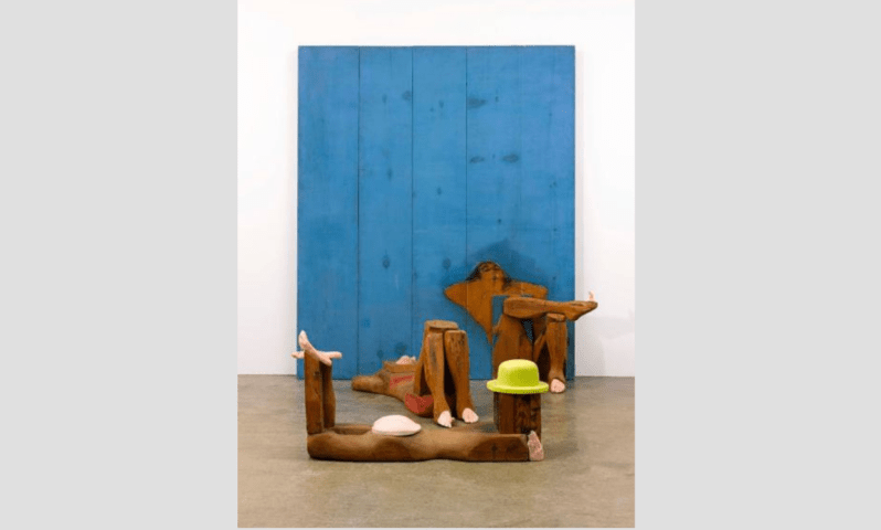 Marisol, The Bathers, 1961-1962, painted wood panel, graphite, plaster cast, and sculpted wood, 84 in. × 70 1/4 in. × 63 in. Promised gift to Crystal Bridges Museum of American Art, Bentonville, Arkansas. Abstract art with wooden figures, exaggerated limbs, blue backdrop, lime green hat on floor.
