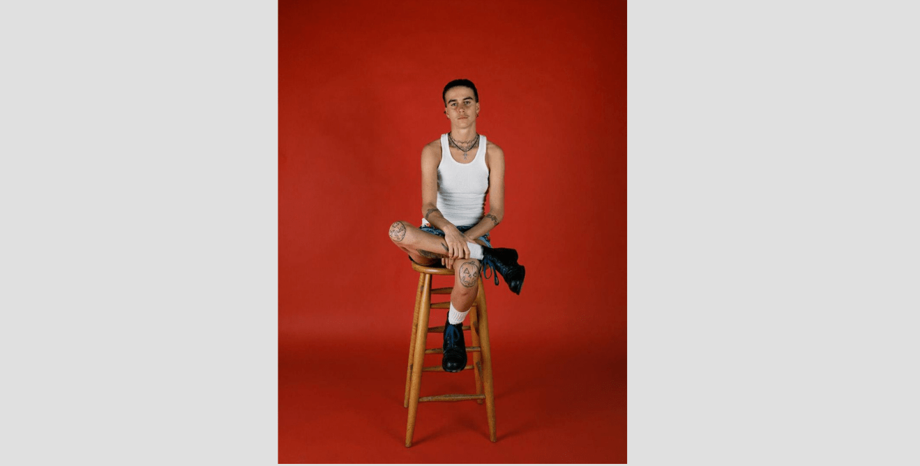 Catherine Opie, Pig Pen, 1993, printed 2013, chromogenic color print, 40 × 30 in. (101.6 × 76.2 cm) Framed: 42 × 32 × 2 in. Crystal Bridges Museum of American Art, Bentonville, Arkansas, 2021.15 © Catherine Opie. Young man in white tank shirt with tattoos on his knees sits on a stool in front of a red background