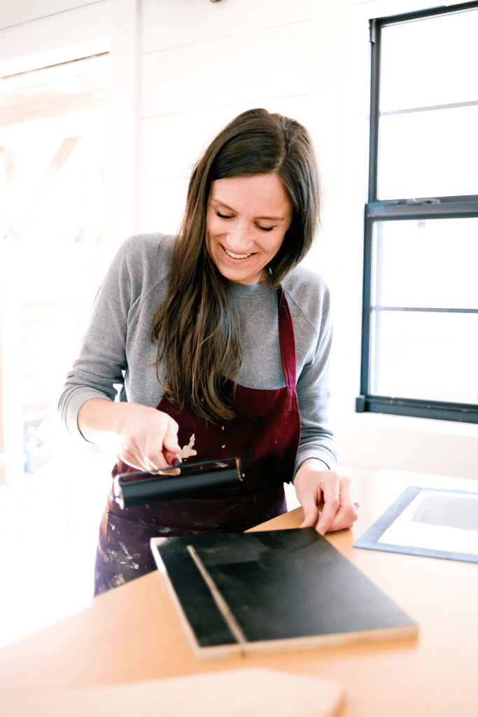 Woman painting wood indoors wearing maroon apron and gray shirt by a window.